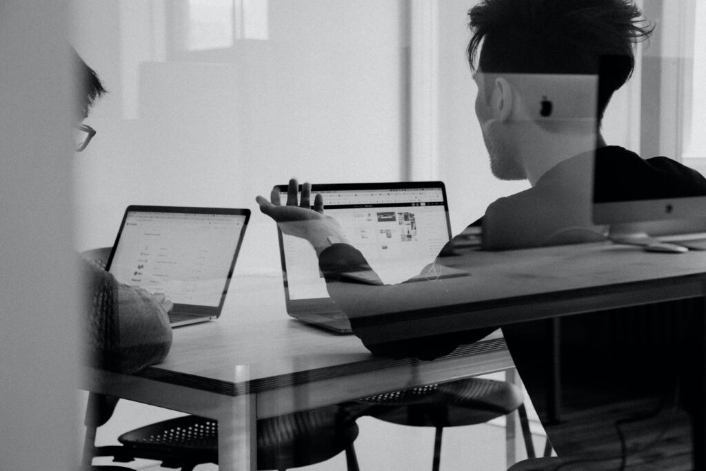 Black and white image of two men with laptops open discussing work.